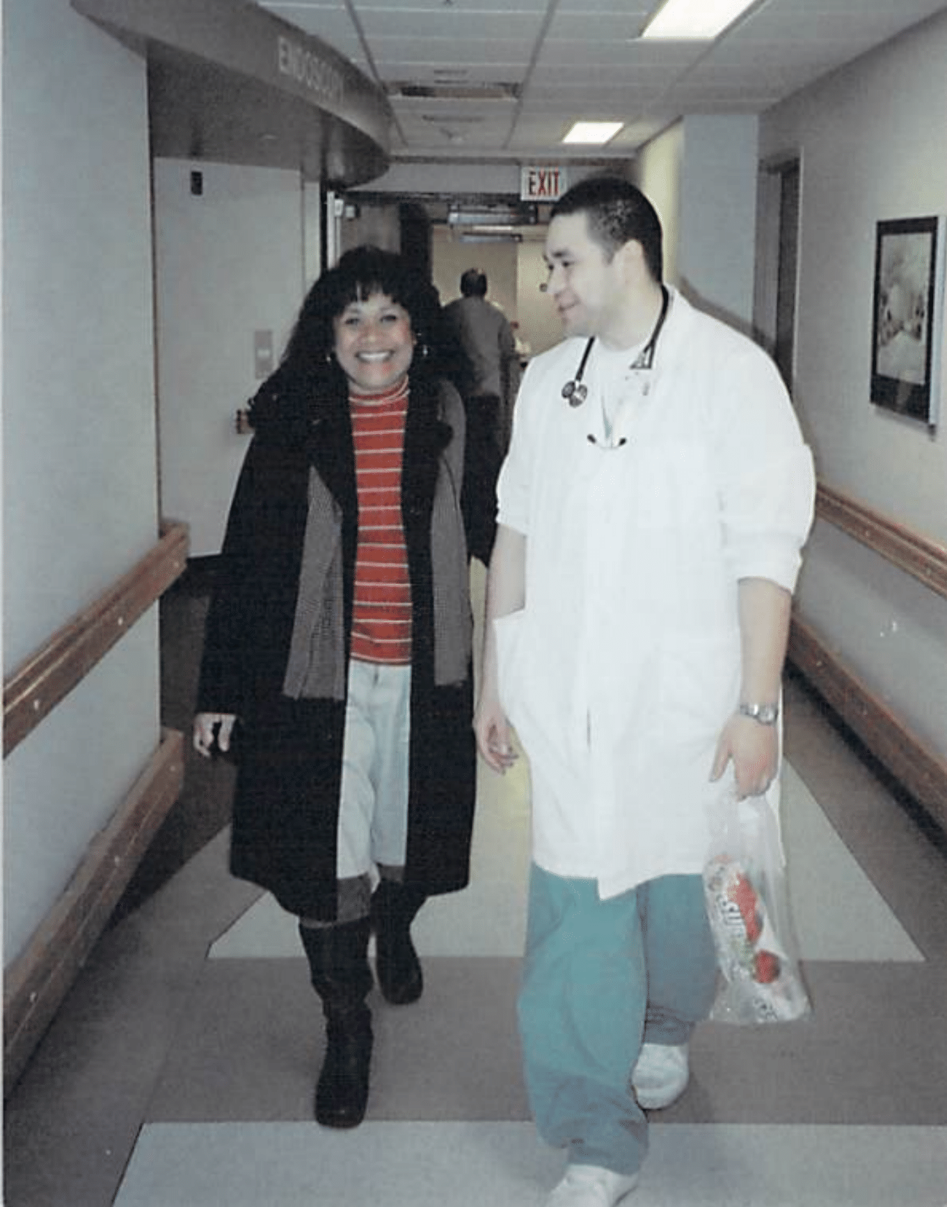A woman with long, dark hair wearing a coat and a red striped sweater is walking down the hall of a hospital with a doctor, wearing a white over coat. He has a stethoscope around his neck and he's smiling, looking at the woman beside him, who is his mother.