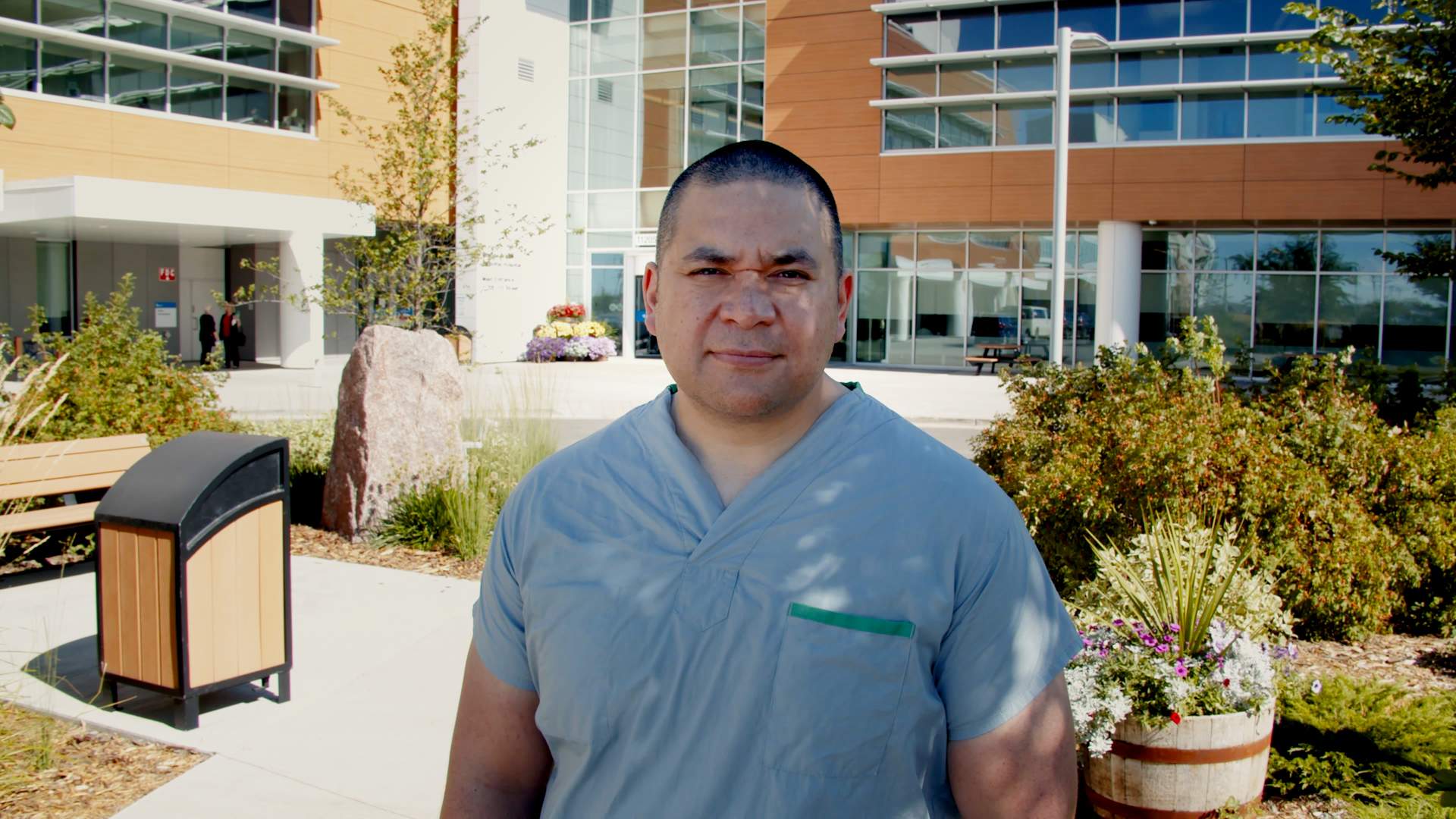 Dr. Alika Lafontaine, a man with closely shaved hair on his head, is standing in blue scrubs, outside of a hospital in Grande Prairie, Alberta.