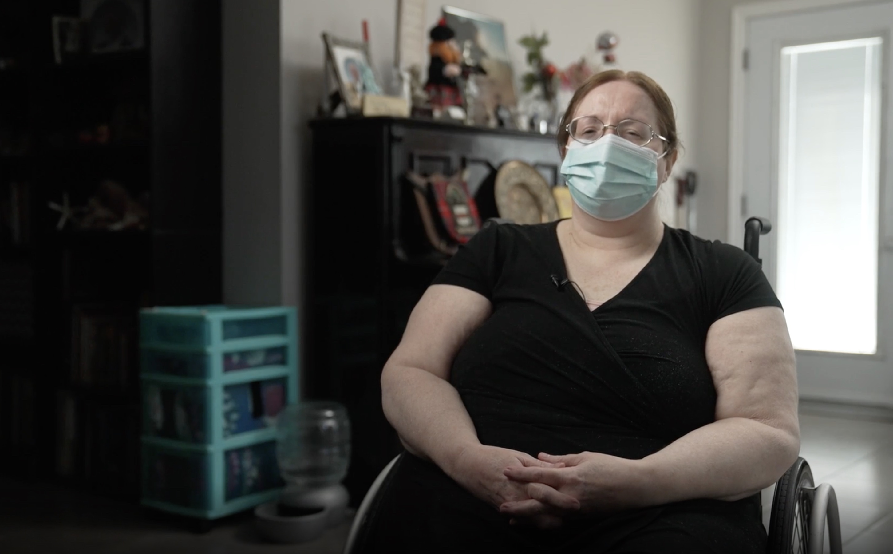 A disabled woman in a medical mask sits in her wheelchair inside her living room.