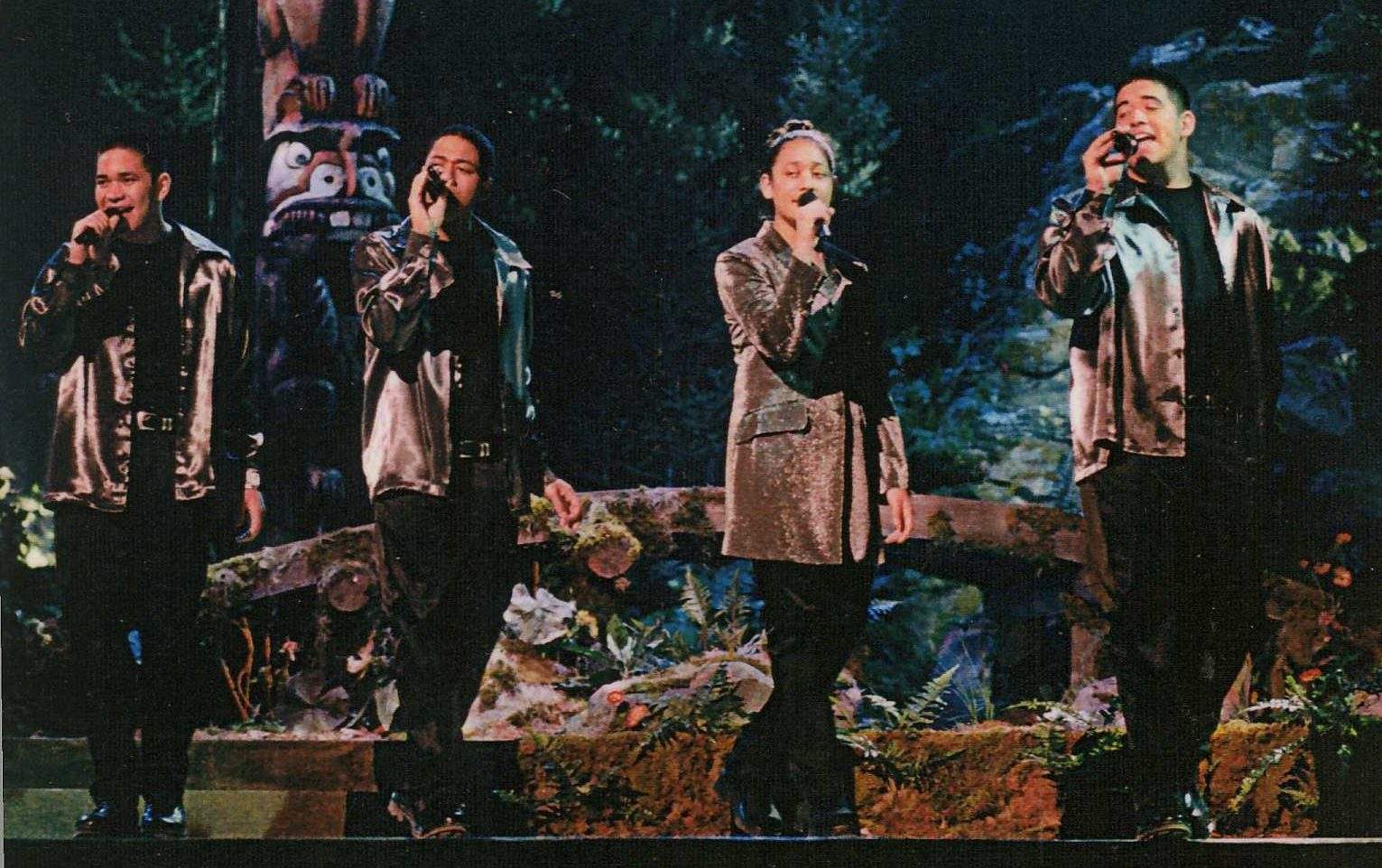 Four members of the pop-group, The 5th Generation, holding wireless microphones while perrming on stage for a live audience at an Indigenous award show in 1999. They are wearing matching outfits of a shiny jacket and black blanks.