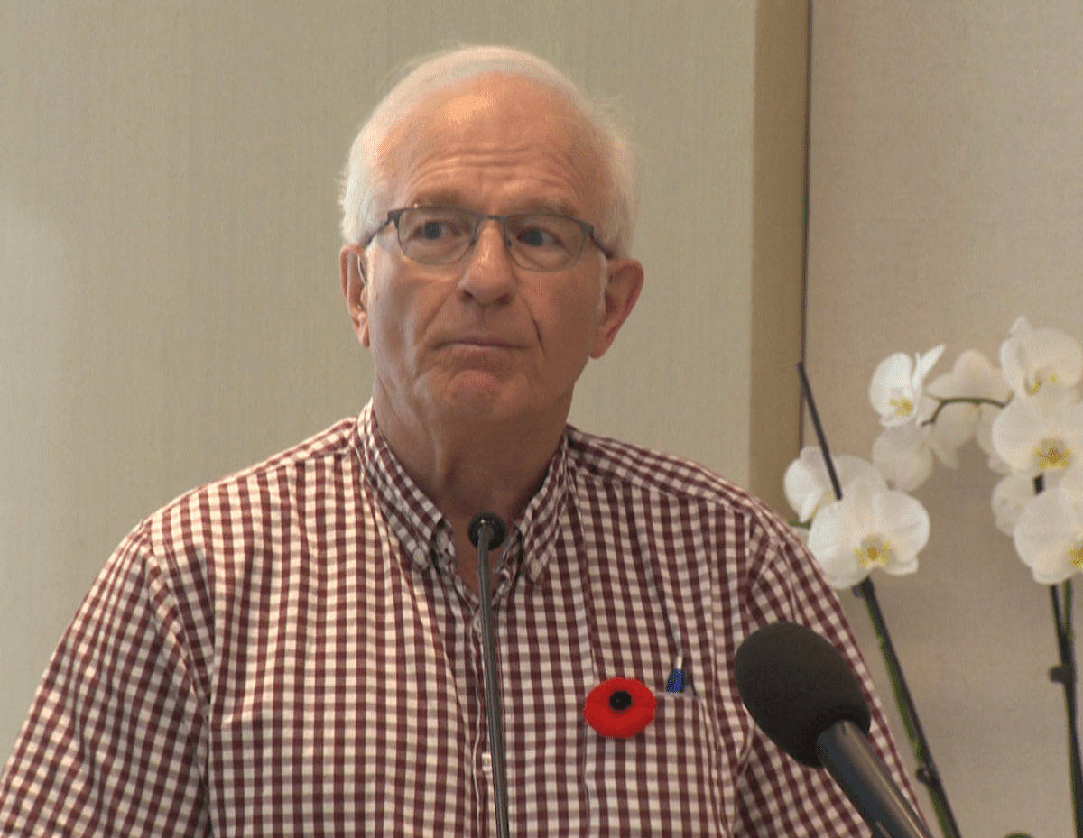 A man wearing glasses and a checkered shirt with a poppy pinned to it stands at a podium.
