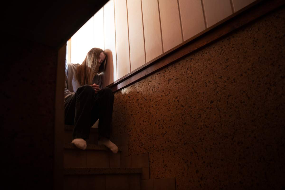 Teenager sitting on indoor steps leading to a low lit cellar and leaning against a wall with an expression of despair.