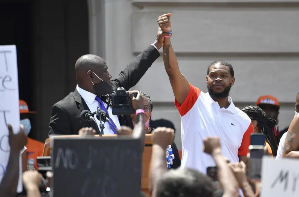 Attorney Benjamin Crump holds up Kenneth Walker's fist in front of a podium.