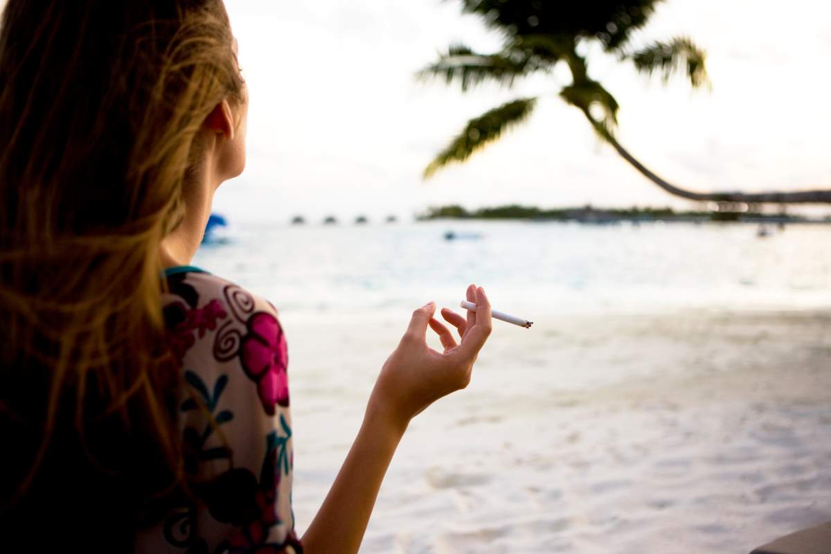 A woman smoking a cigarette on the beach. There is a palm tree in the background.