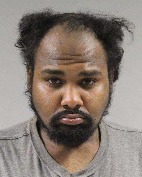 Mabanza is pictured in a mug shot with a neutral expression on his face. He has a dark complexion, with black curly hair and a full beard that is trimmed close to his face. His hairline is receding. He is wearing a beige shirt.