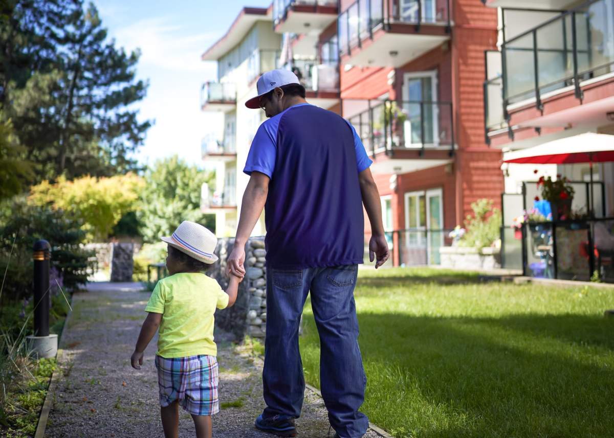 A man holds a young boys hand as they walk down a path behind an apartment building.