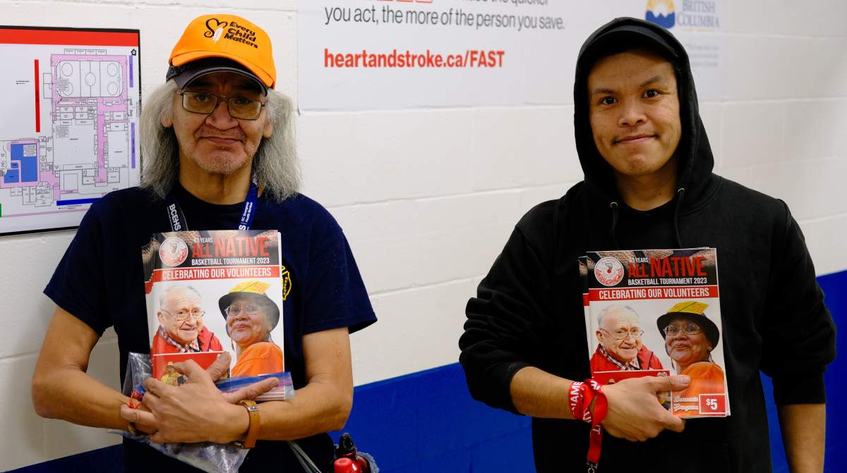 Two people stand holding booklets for the All Native Tournament in front of them.