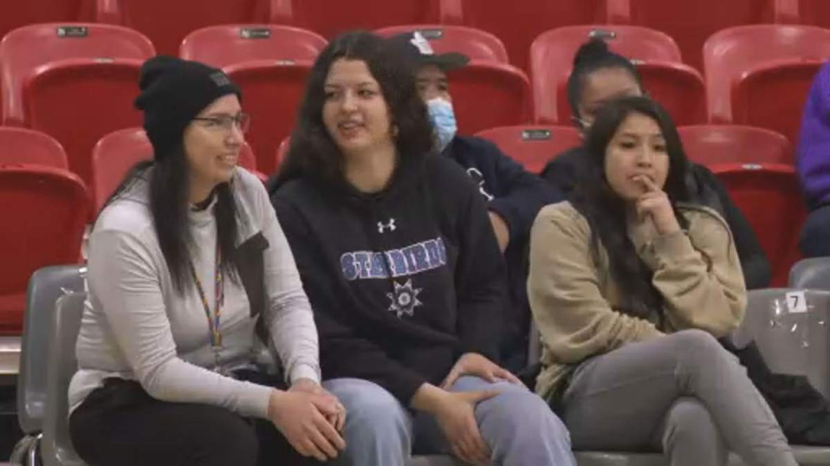 Three women sit next to eachother on a bench watching a basketball game.