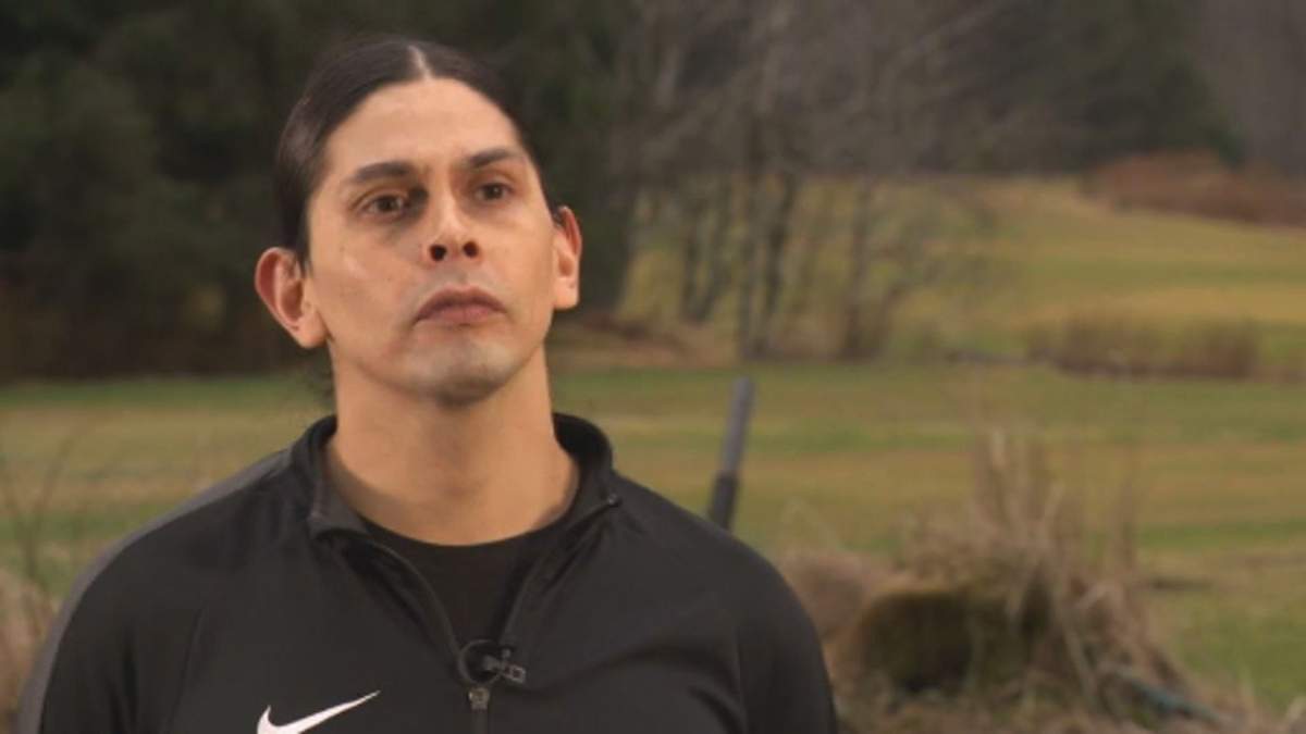 A man stands looking at the camera. There are trees in the background, he's wearing a black sweater.