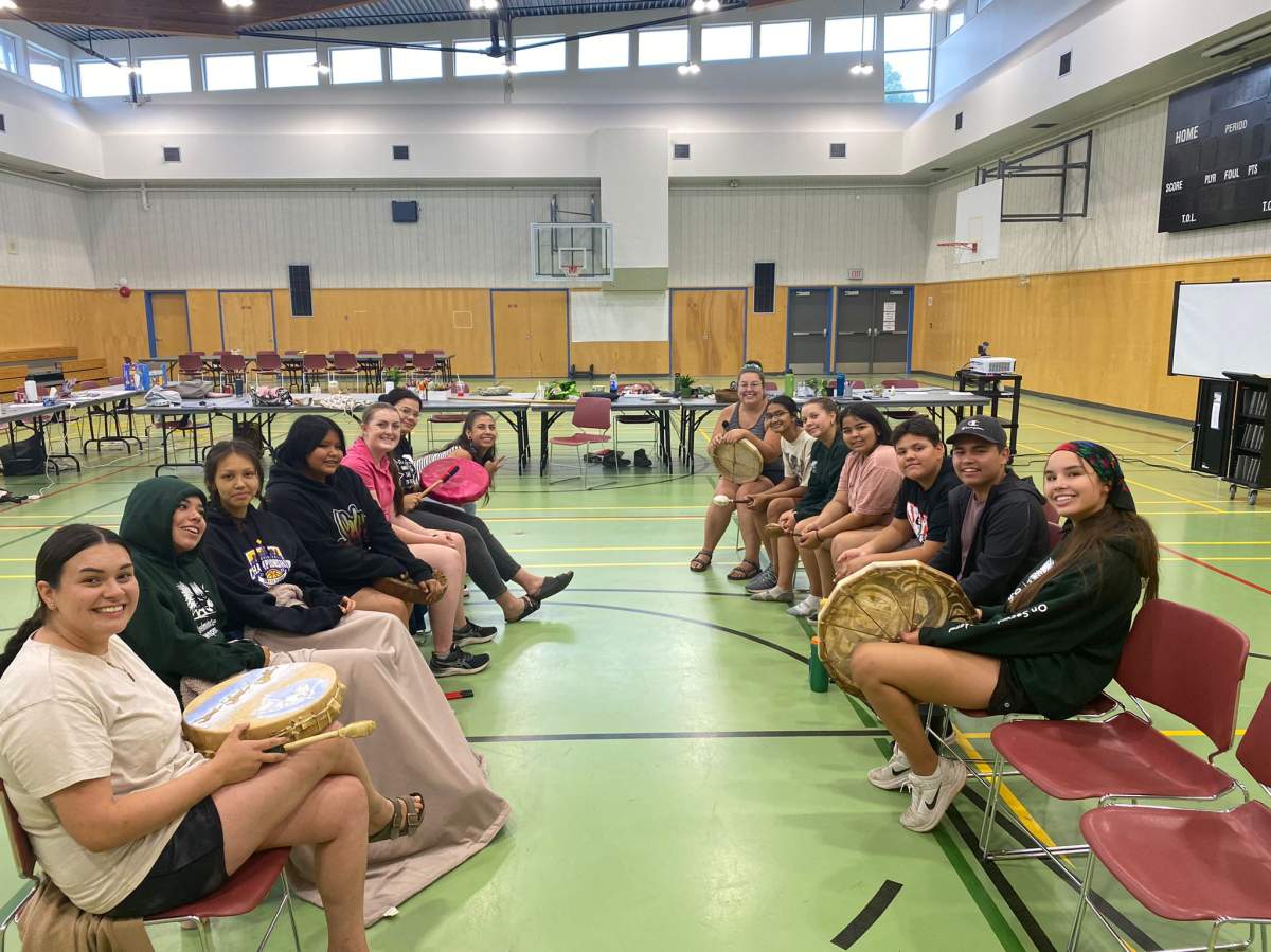 A group of Indigenous youth sit on chairs facing one another. They're in an older gymnasium, the floor is green.