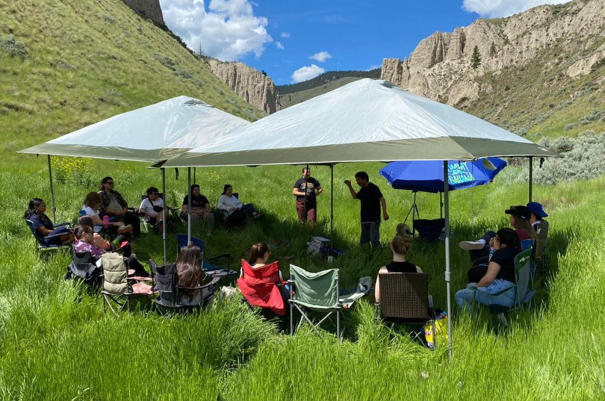 Two tents sit atop green grass as a group of people sit on chairs underneath them. In the background you can see a mountain and blue skies.