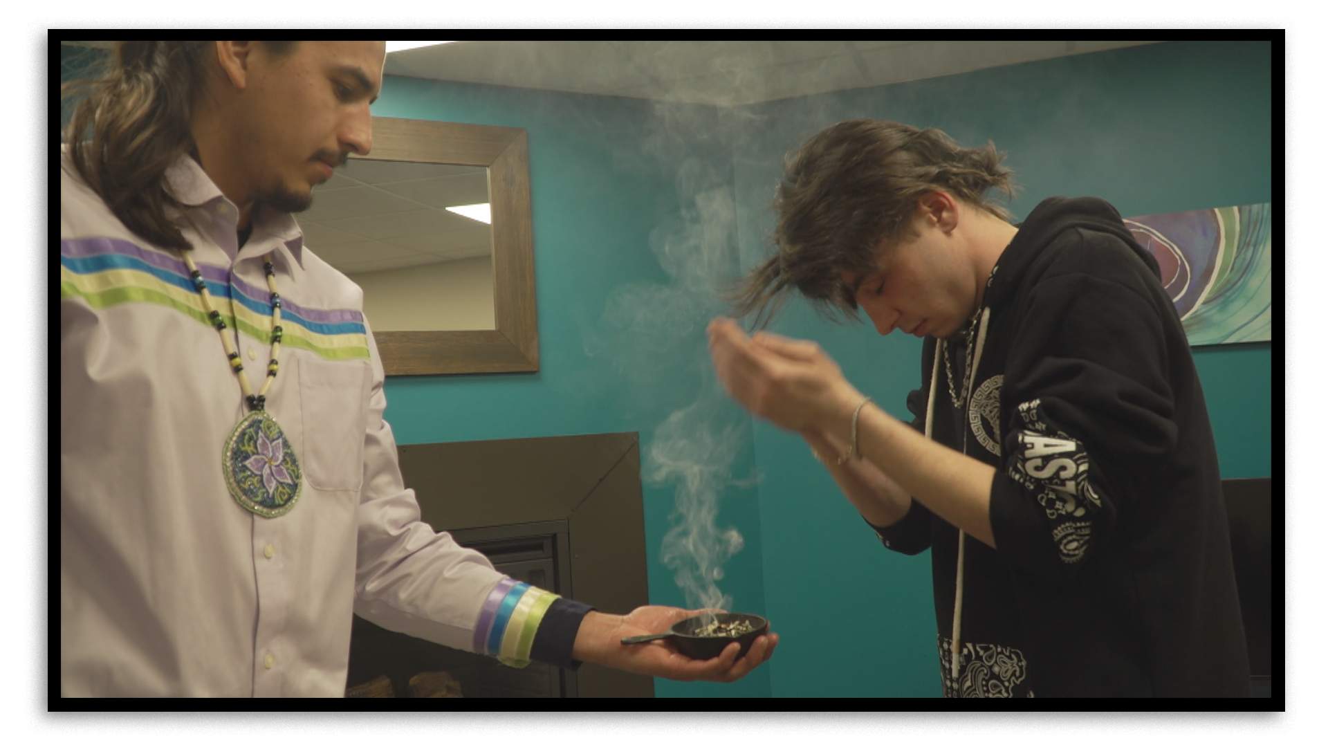 Two men stand in a room whose wall colour is teal. On the left, a man wearing a white shirt with long brown hair, a goatie and a traditional beaded necklace holds a small bowl of burning herbs in his outstretched left hand. A wisp of smoke rises from the bowl and the 21-year-old man on the right, Ethan Pokno, cups his hands to capture it, pulling some of the smoke towards himself in a ceremony known as smudging. He’s wearing a black sweatshirt with white logos on it, a friendship bracelet on his left wrist, and a chain necklace.