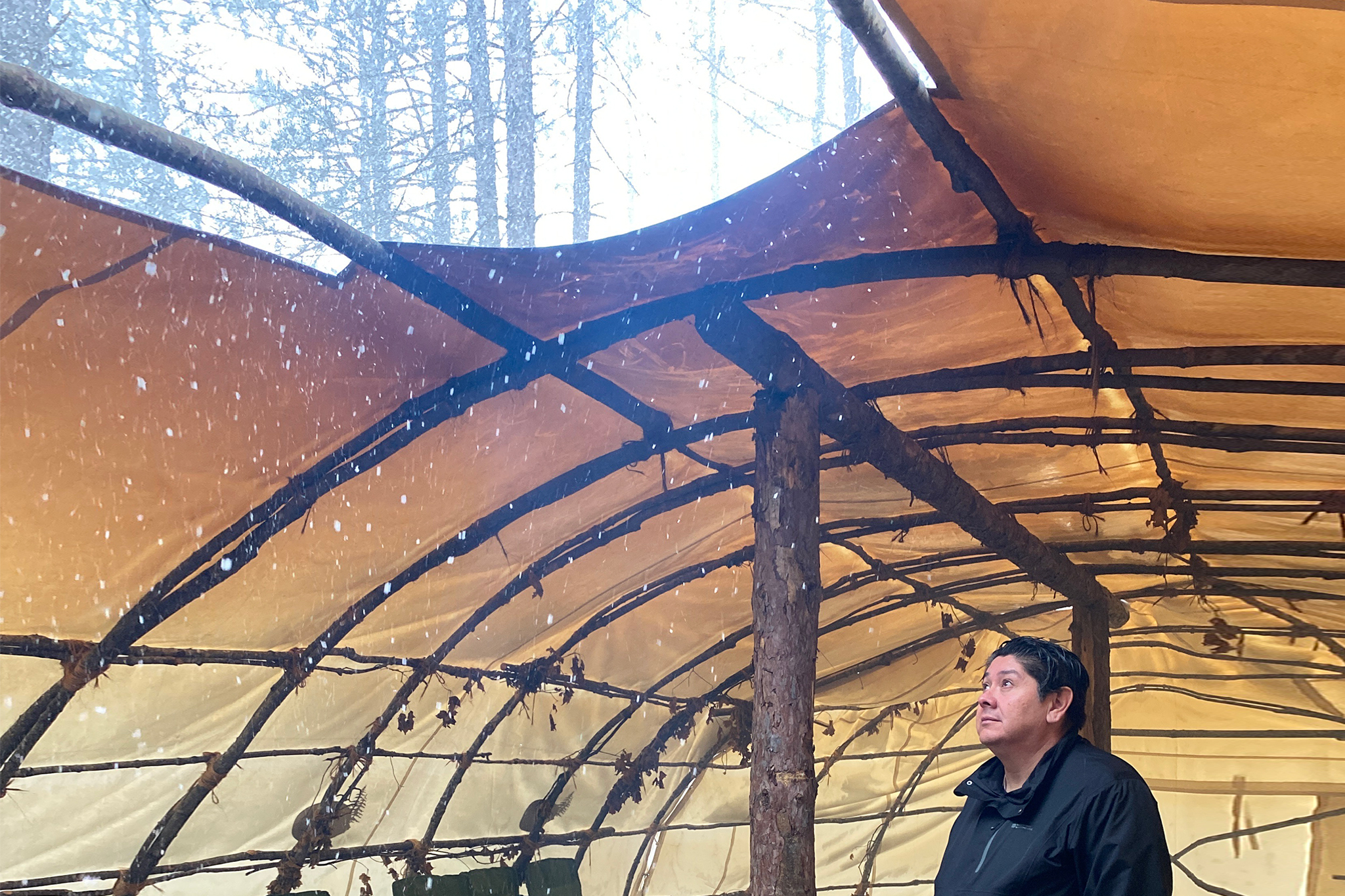CEO Kerry Francis stands inside a teaching lodge warmed by a fire at his feet. He looks up through an opening in the lodge's canvas roof that acts as an air vent. Smoke arises through the opening while snowflakes drift in through it.