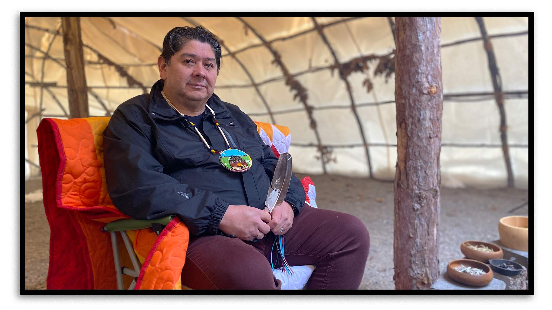 Kerry Francis, Nogdawindamin’s CEO, sits in a camping chair covered in an orange blanket inside the organization’s teaching lodge. Francis has black, slighting greying hair and is wearing a dark blue jacket, maroon-coloured pants and a necklace in a traditional First Nations style. He holds a feather in his hands. The sides and roof of the teaching lodge are made of bent saplings, with sturdy tree trunks holding up the centre. The lodge is covered in white canvas.