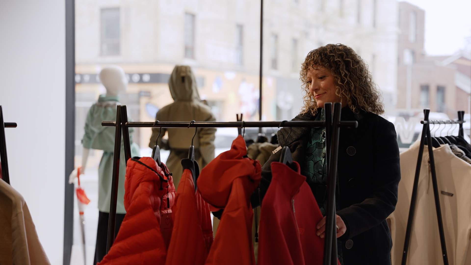 Kelly Drennan, a woman with blond and brown curly hard, is looking through jackets on a rack inside a clothing store.