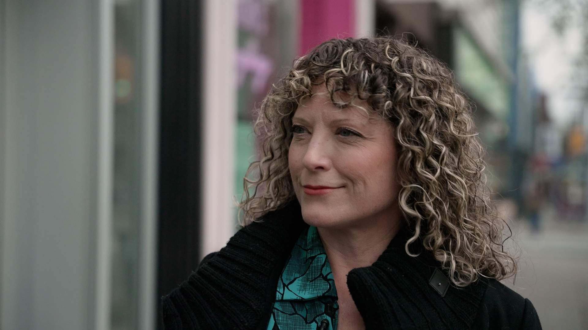 Kelly Drennan is the Founding Executive Director of Fashion Takes Action, a Canadian non-profit. She has dirty-blonde, curly hair and is looking away from the camera while walking down the street. She is wearing a black coat over a green dress.