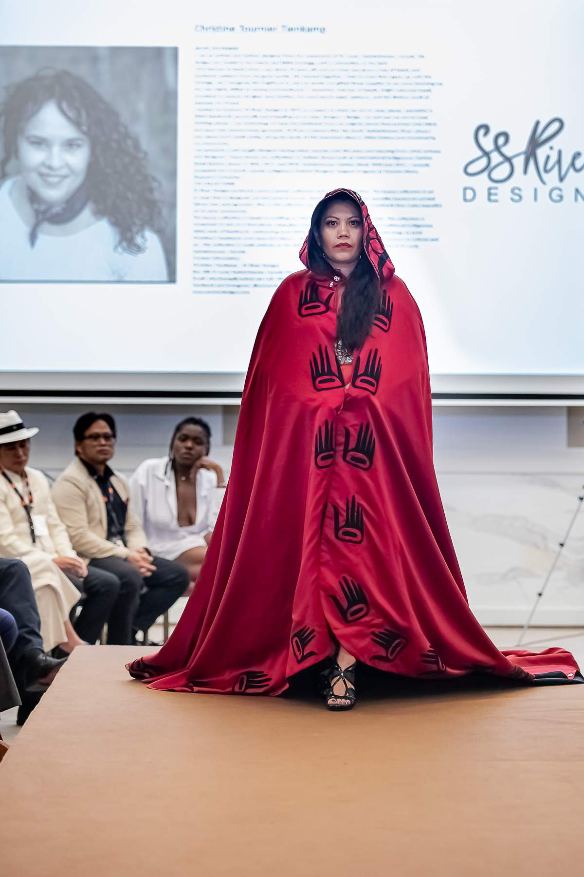 A woman walks down a runway wearing a red cape.