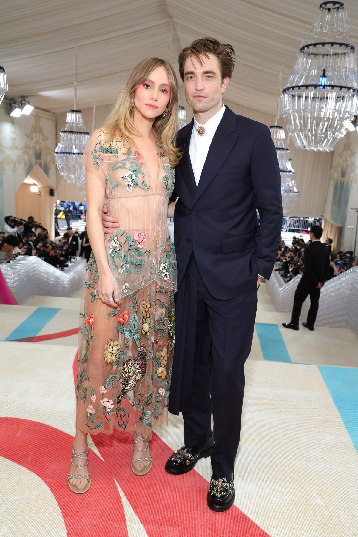 Suki Waterhouse wears a sheer, floral adorned full-length gown. Robert Pattinson wears a full suit with a train behind his legs.