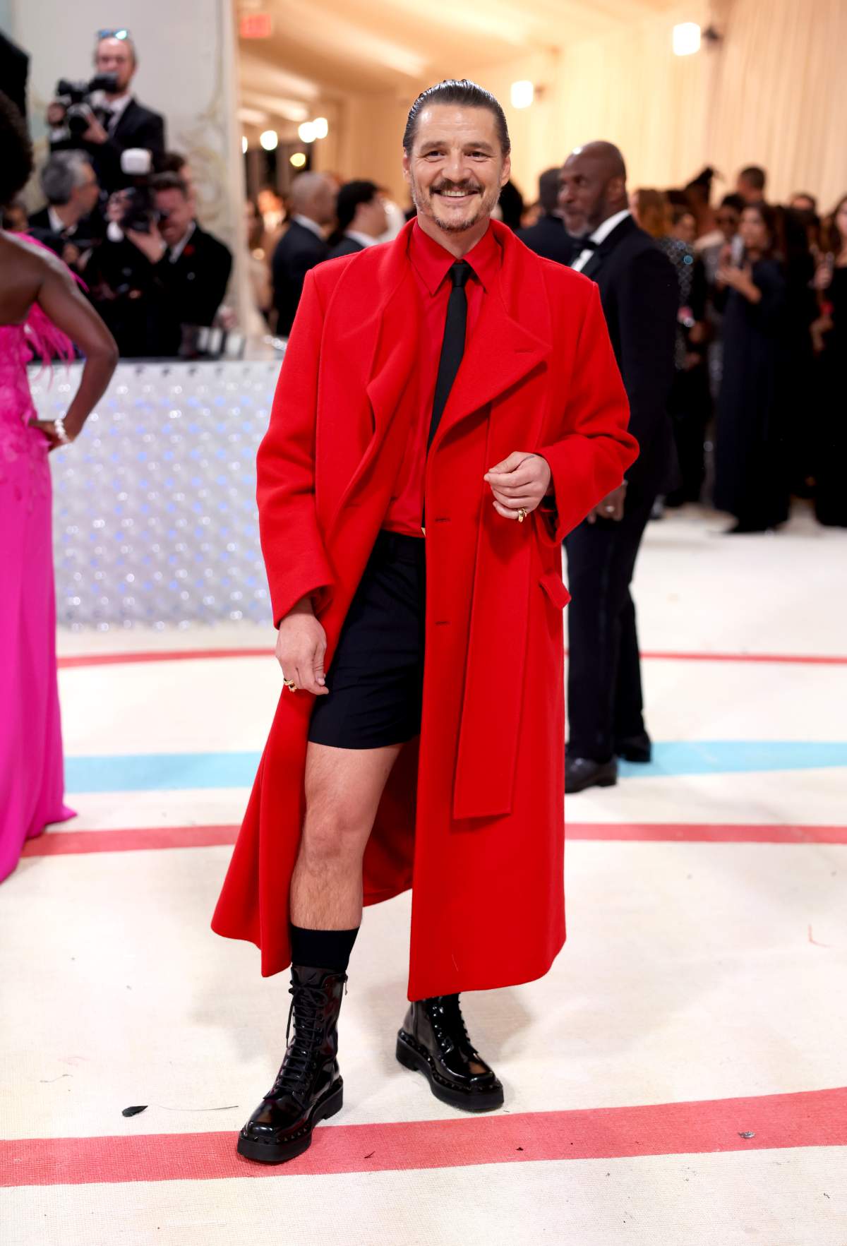 Pedro Pascal in a long red coat, red shirt, black tie and black shorts. He is wearing long black boots.