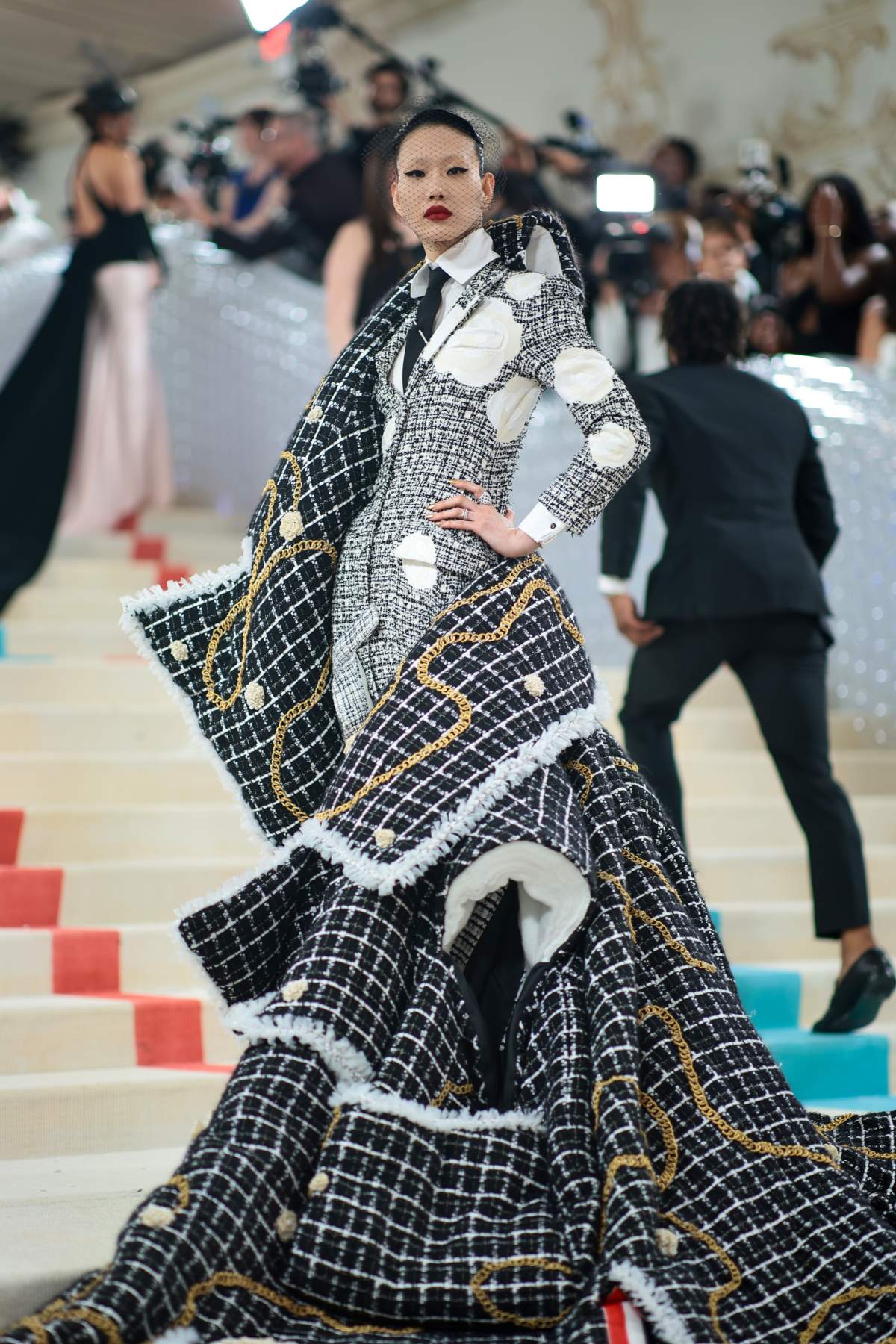 Sora Choi in a tweed suit with white spots. She is wearing a heavily structured cape made to look like an overthrown jacket, adorned with gold chain.
