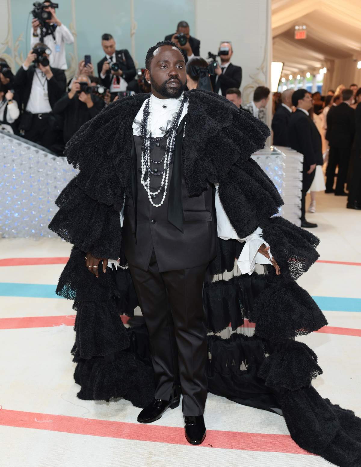 Brian Tyree Henry wearing a stylish black suit, long pearls and a large, frilly cape.
