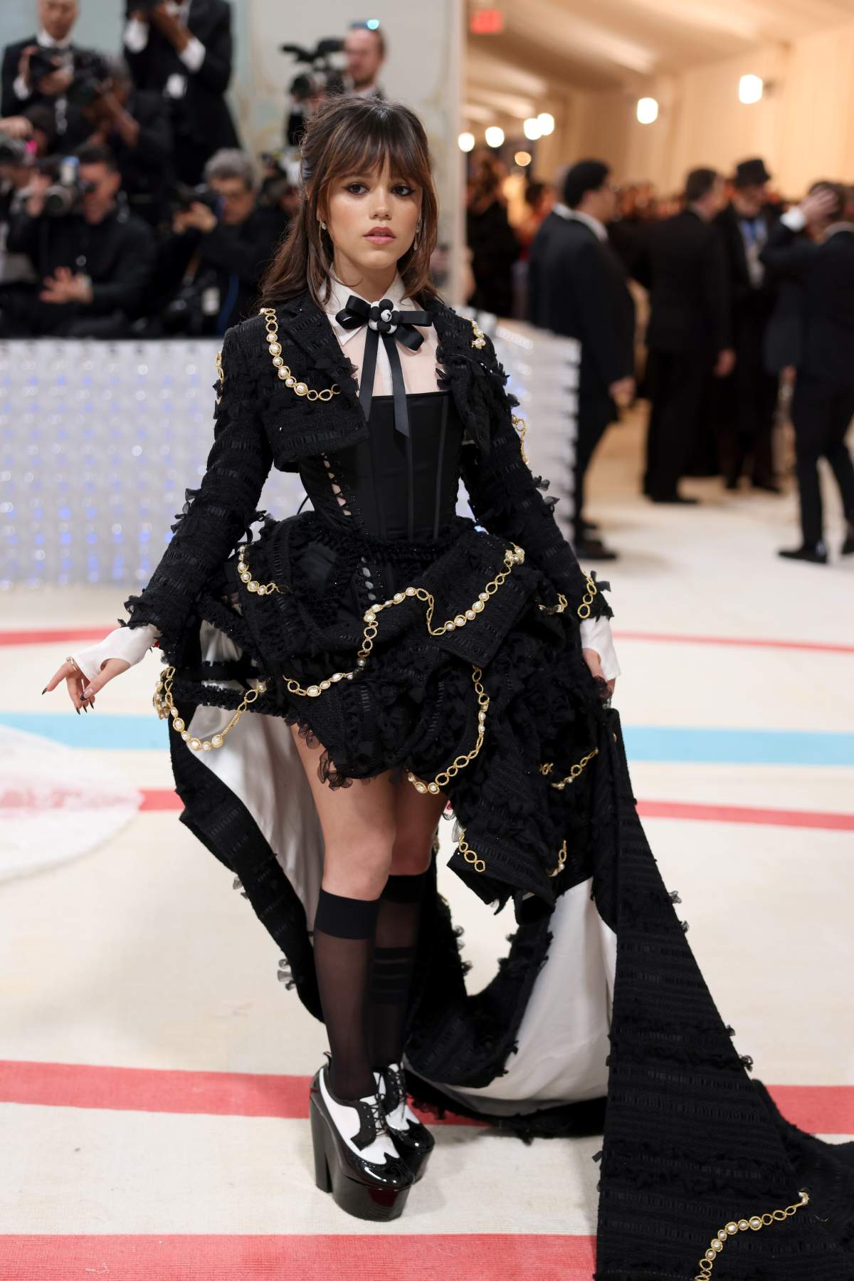 Jenna Ortega in a high-low black gown with frills and gold chain. She wears a black bowtie.
