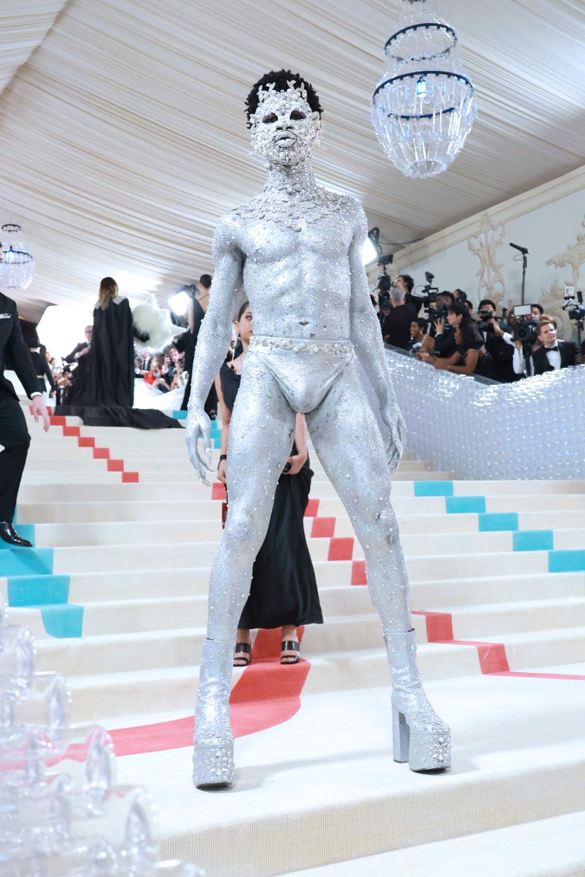 Lil Nas X in silver body paint. He wears high heeled boots, underwear and a lavish mask made from silver gems and pearls.