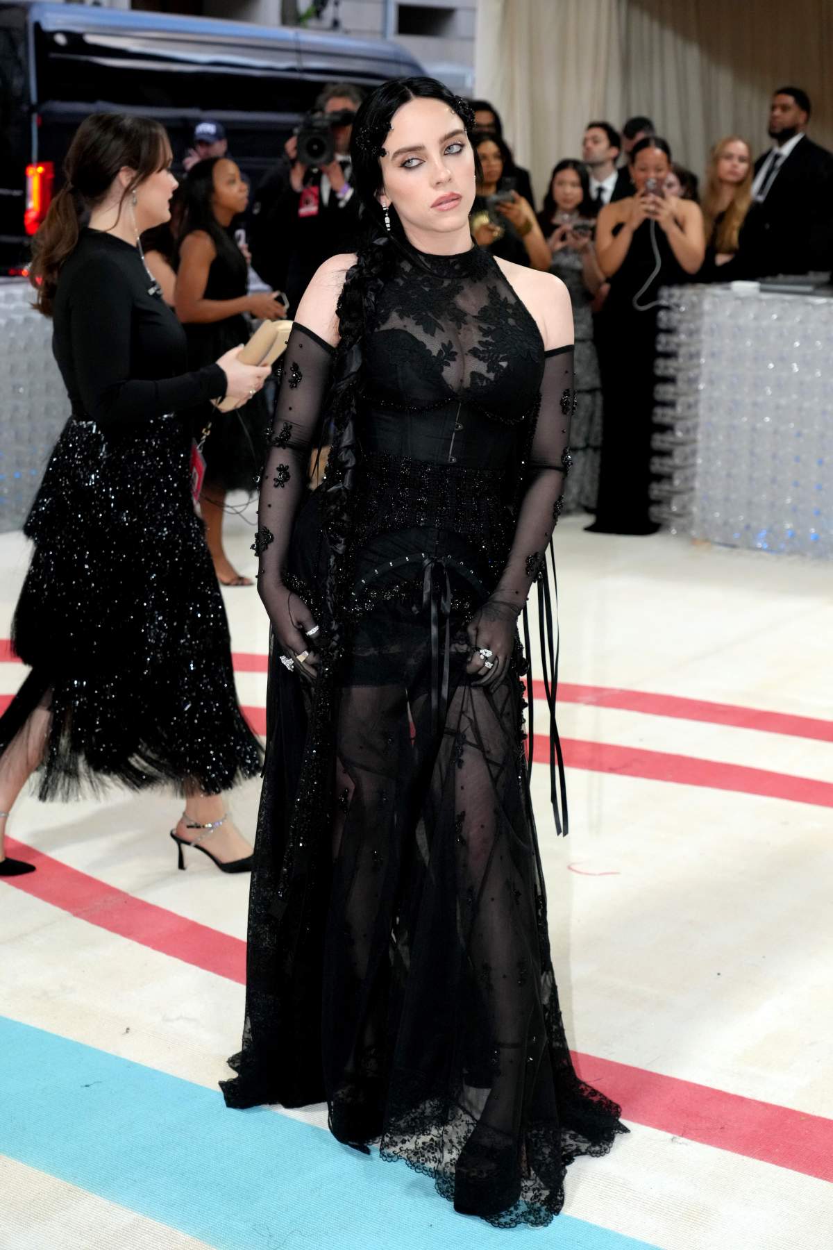 Billie Eilish in a full-length black, sheer gown. Her hair is pleated and pressed, and she wears long, sheer black gloves.