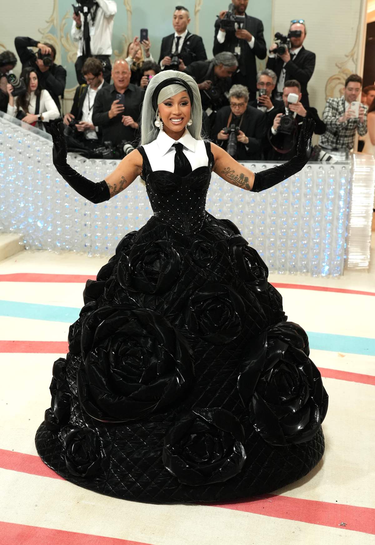 Cardi B in a stiff, leather-looking ball gown adorned with large, faux roses. Her hair is silver and teased high with a headband.