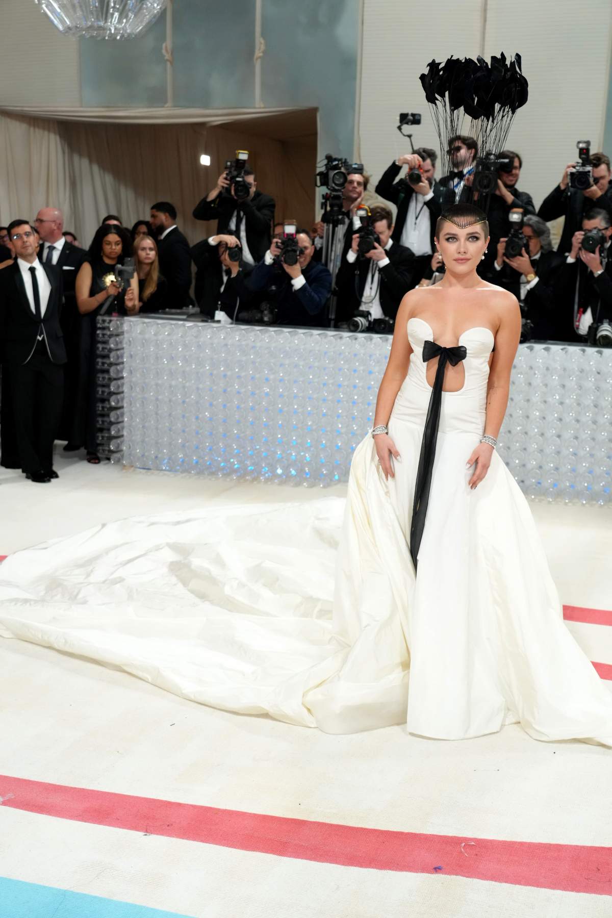 Florence Pugh in a full-length white gown with a black bow and large train. Her hair is buzzed, and she wears a tall crown of feathers.