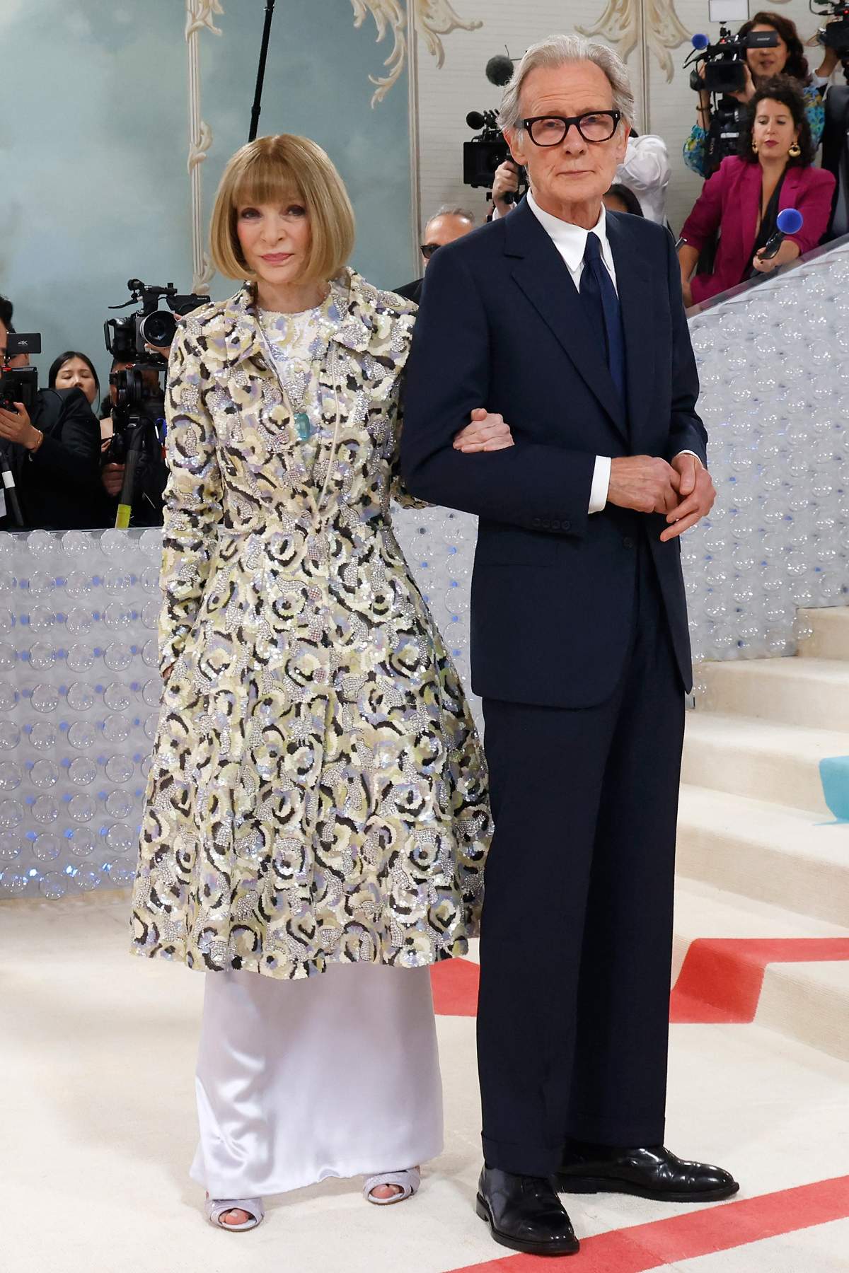 Anna Wintour wears a full-length white gown under a long gold and silver coat. Bill Nighy wears a classic suit.