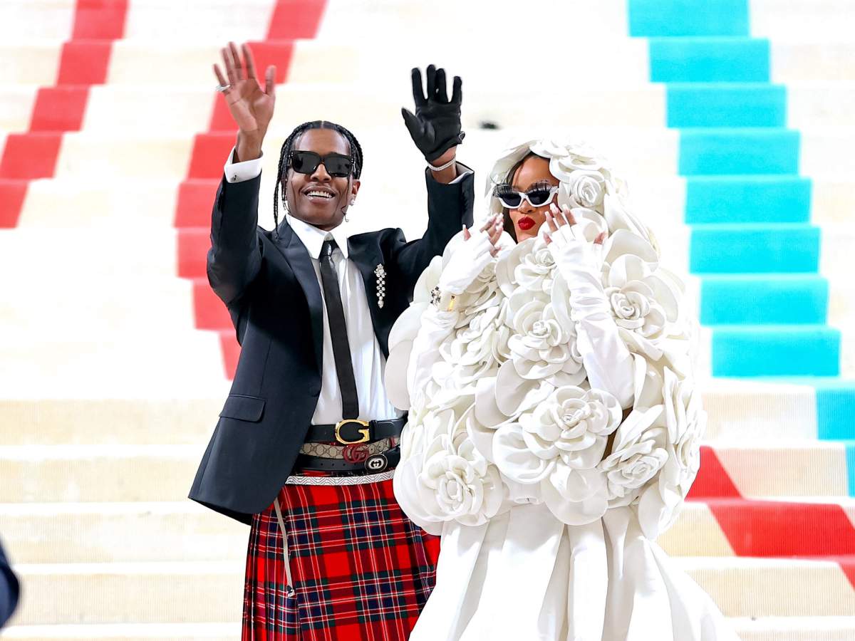 Rihanna wears a white gown topped with a hooded jacket of white roses. A$AP Rocky wears a red kilt, white button up, black tie and a black jacket. They both are wearing black sunglasses.
