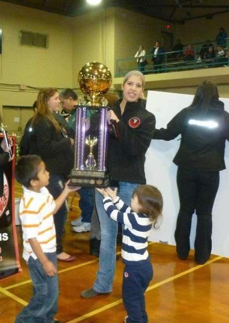 A woman stands holding a large trophy with two children in front of her reaching for the trophy.