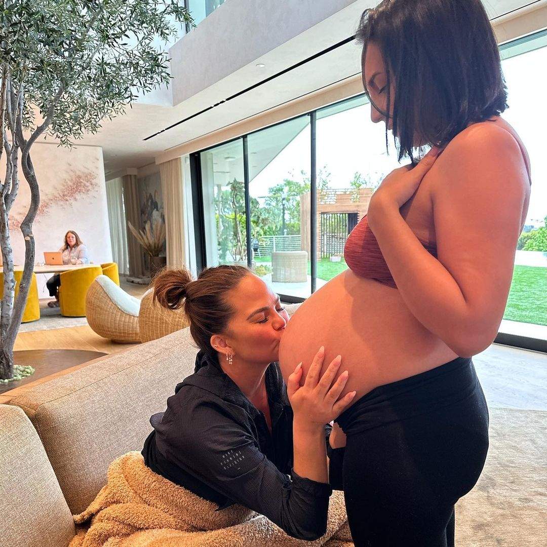 Photo of Chrissy Teigen kissing her surrogate's baby bump before the birth of her fourth child, Wren Alexander Stephens.