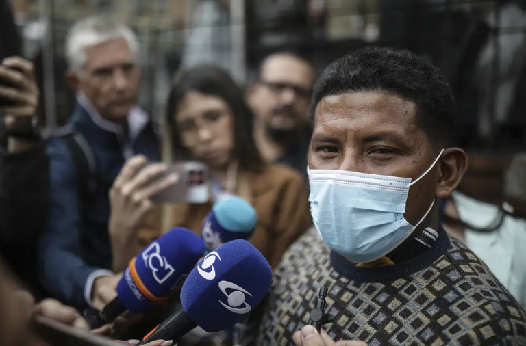 Manuel Ranoque wearing a blue medical mask. He is speaking to reporters with microphones.