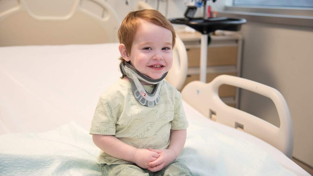 A toddler in a neck brace smiles while sitting on a hospital bed.