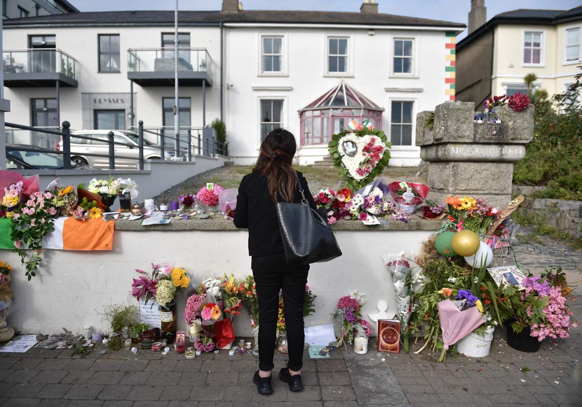 A mourner stands outside Sinéad O'Connor's home. There are flowers and gifts surrounding the gate.