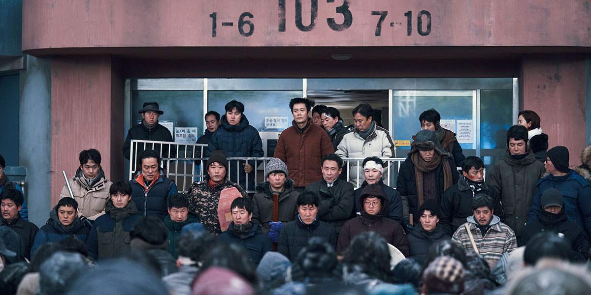 A still from 'Concrete Utopia.' People stand in coats in front of a shabby looking building.