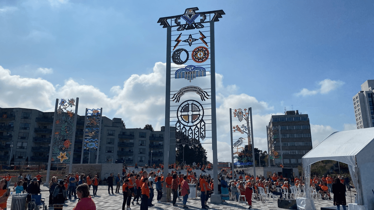 Five tall panels of artwork made up of thousands of multi-coloured glass beads stand at the base of James Street North on a sunny day, while people in orange shirts gather below the structures.