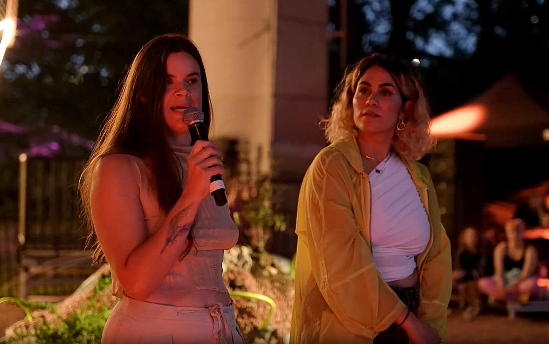 Larissa Belcic stands with long dark hair, holding a wireless microphone looking towards a crowd of people. She's wearing a light coloured short-sleeve t-shirt. Beside her is Michelle Shofet, who has cropped curly hair and is looking towards Larissa as she speaks.