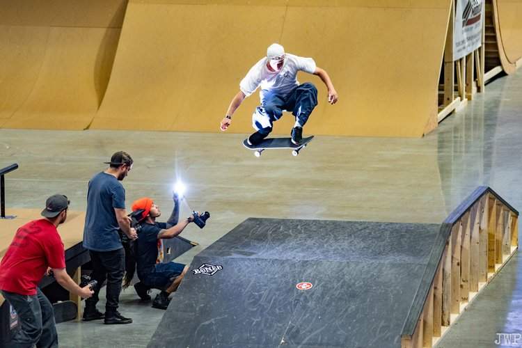 A person does a skateboard trick on a ramp, he's being photographed by a bunch of people.