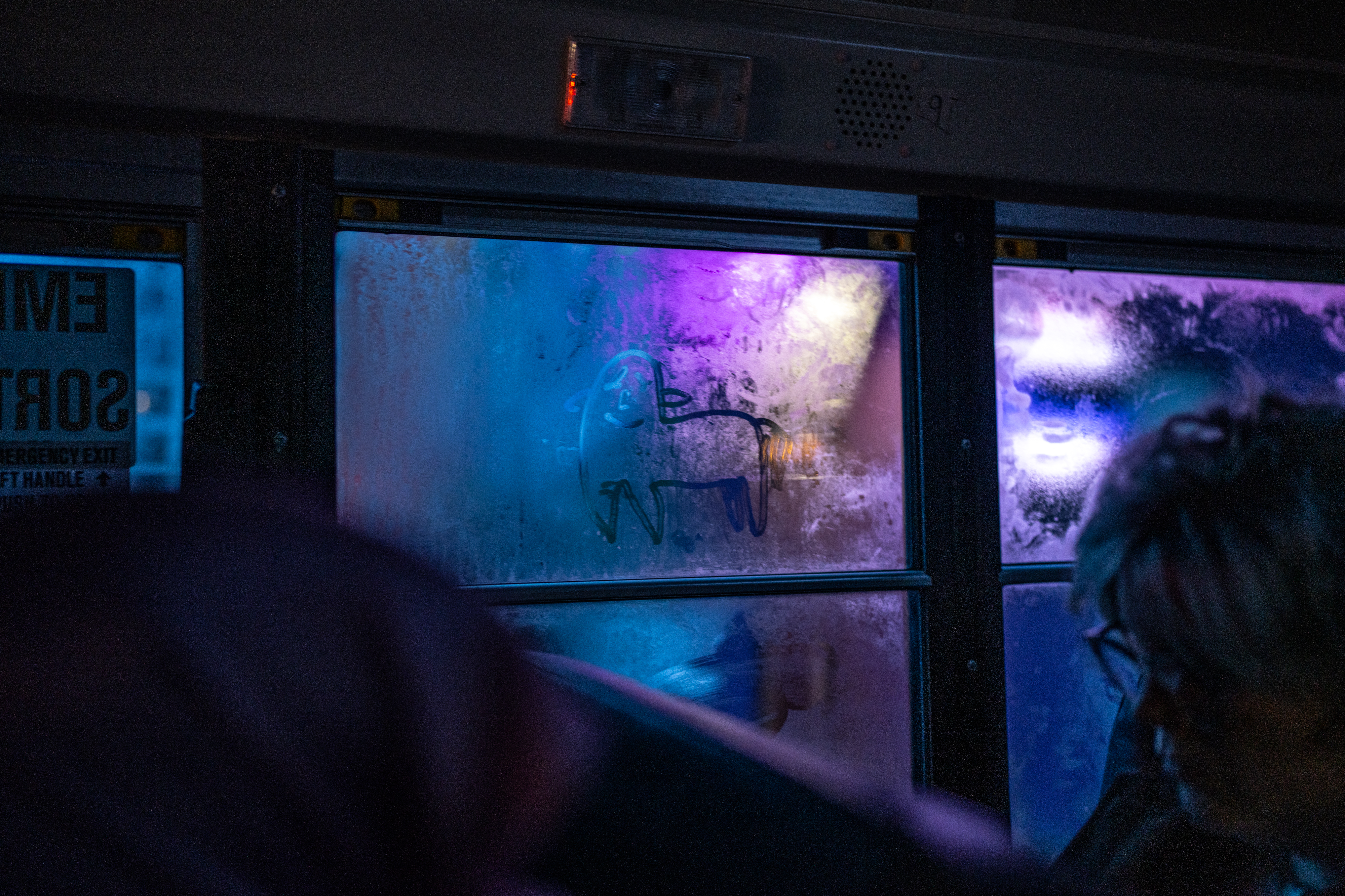 A foggy school bus window is lit by blue and purple light.