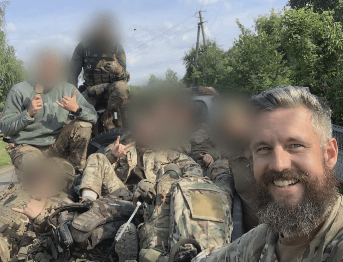 A group of soldiers smile for a selfie while seated in a pickup truck in Ukraine. The faces of all but one - Canadian Dave Smith - are blurred.