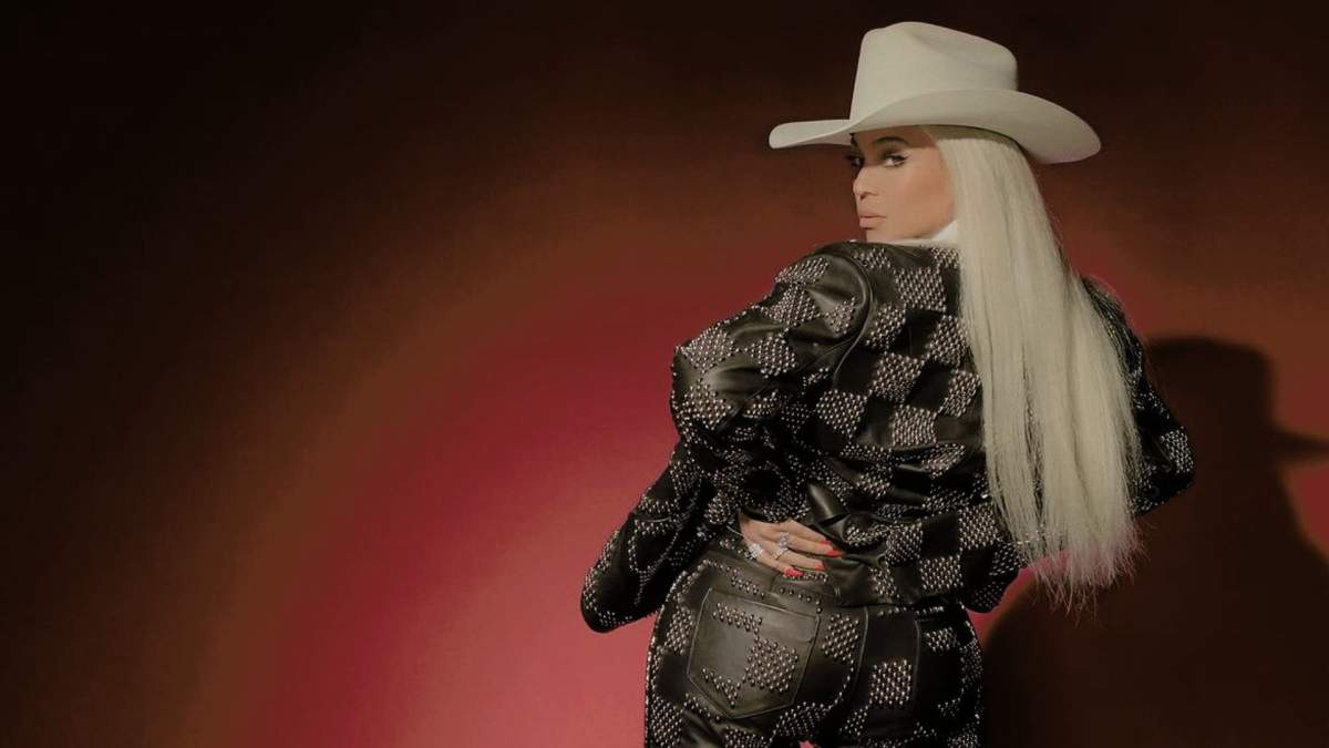 Beyoncé in a cowboy hat. Her hair is long and blonde. She is wearing a checkered jacket and black pants.