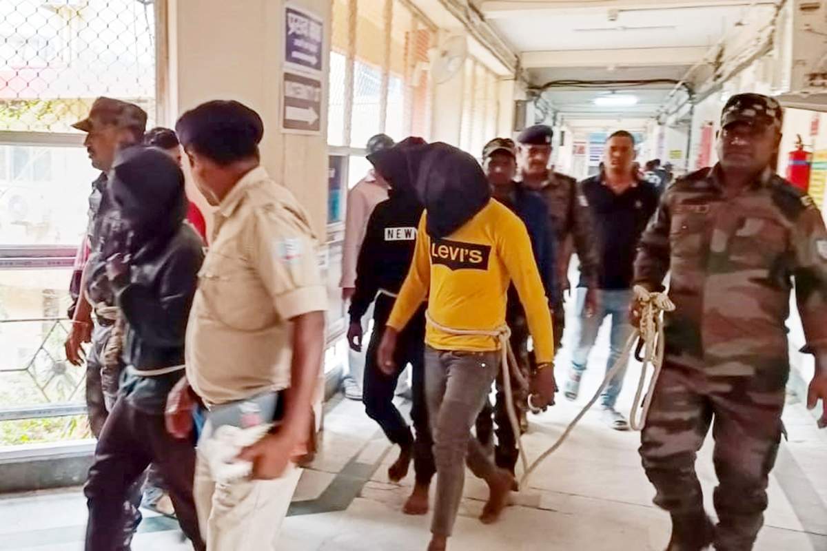 Uniformed police escort three men through a hallway. The three men are wearing casual clothing, ropes around their waists (held by police like a leash), and black bags over their heads.