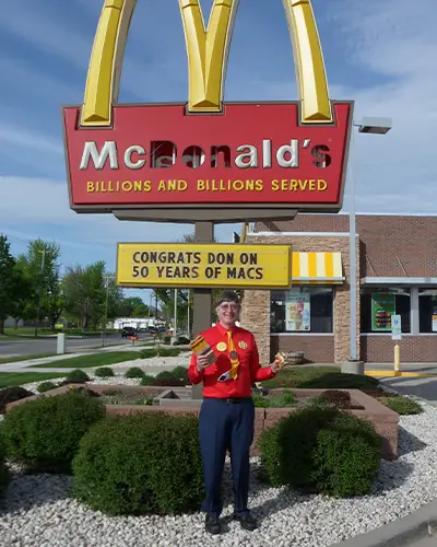 Donald Gorske in front of a McDonald's sign missing the letters D, O, N and S. The menu board below reads, "Congrats Don on 50 years of Macs."