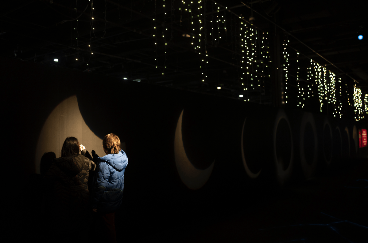 Two kids play in front of a partial eclipse-shaped light on a black wall.