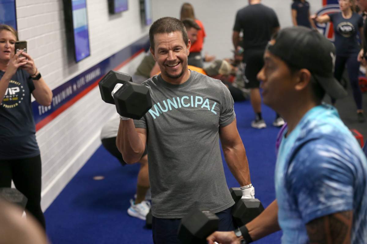 Mark Wahlbeg, in a 'MUNICIPAL' tee, curling a dumbbell inside an F45 gym.