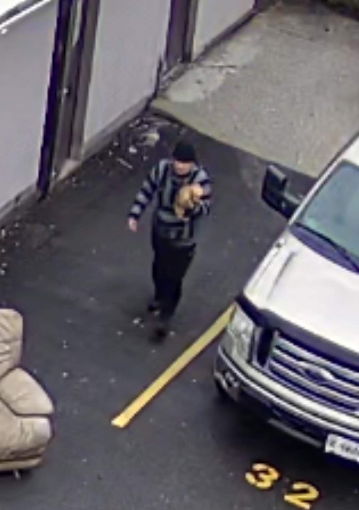 Surveillance image of a man holding a small dog in one arm in a parking lot.