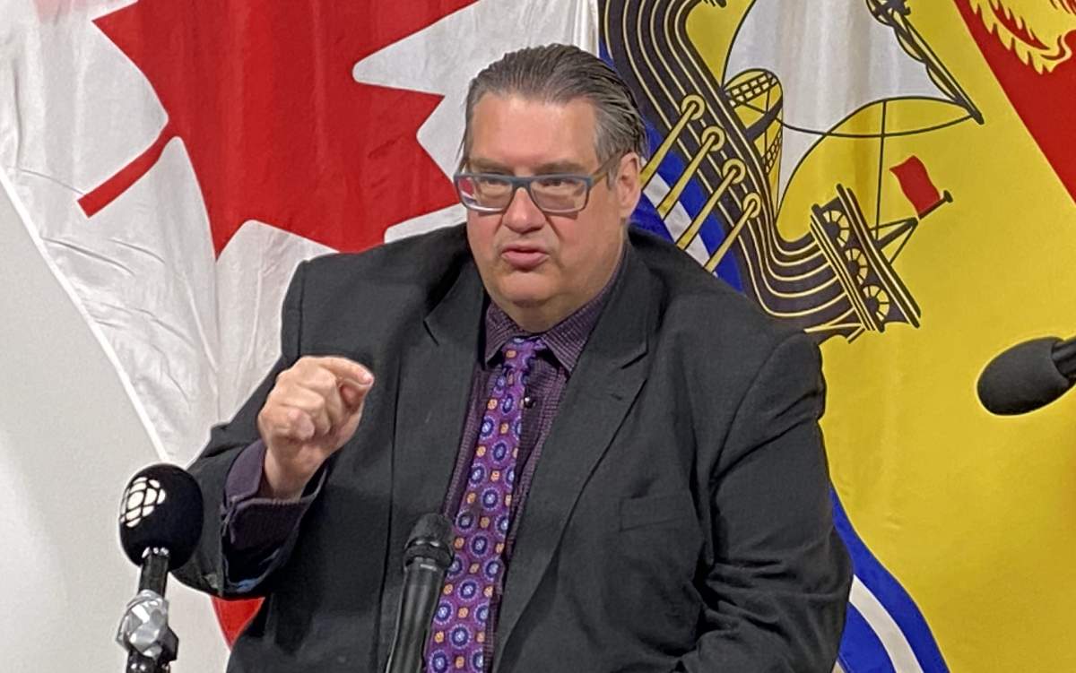 A man with a black suit and a purple tie has his hand raised. He sits in front of a Canadian and New Brunswick flag.